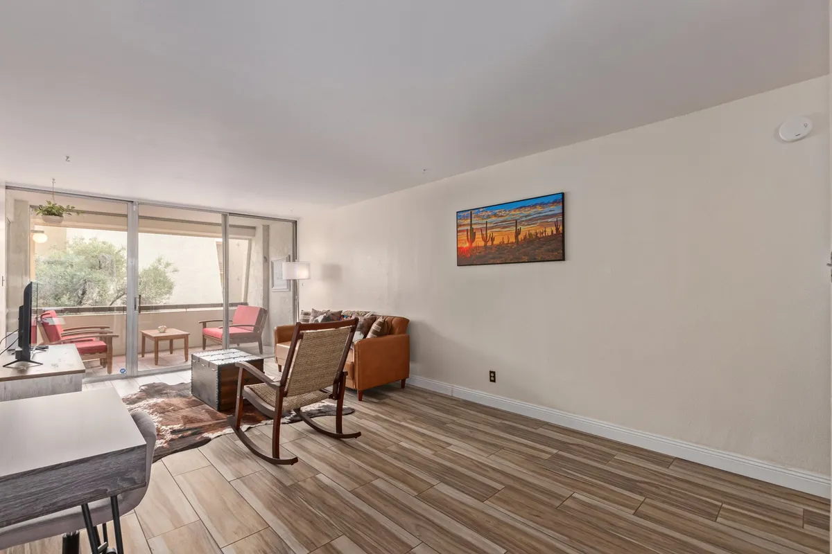 This image features a cozy living room with a sofa, rocking chair, TV, painting, and large windows. The generous natural light suggests a connection to outdoor views—possibly a balcony or sunset scene—while the arrangement invites relaxation and socializing.