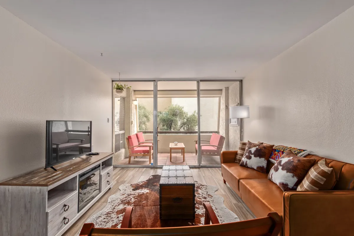 This image features a stylish living area with a leather couch, a wooden coffee table, a television, and sliding glass doors that open up to an outdoor seating space. Abundant natural light pours in, and the decor is modern yet cozy, with elegant touches throughout.