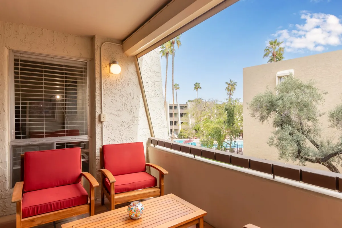 An inviting exterior shot of a balcony or patio area with red chairs and a wooden table, set against a backdrop of palm trees. The setup suggests a space for sipping drinks, relaxing, or sharing a quiet moment while enjoying the view.