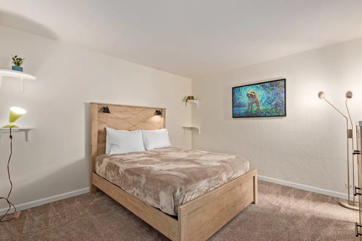 A bedroom with plush bedding, colorful artwork above the bed, a modern floor lamp, and shelves adorned with green plants. The space feels cozy, with soft textures and calming colors that invite relaxation and sleep.