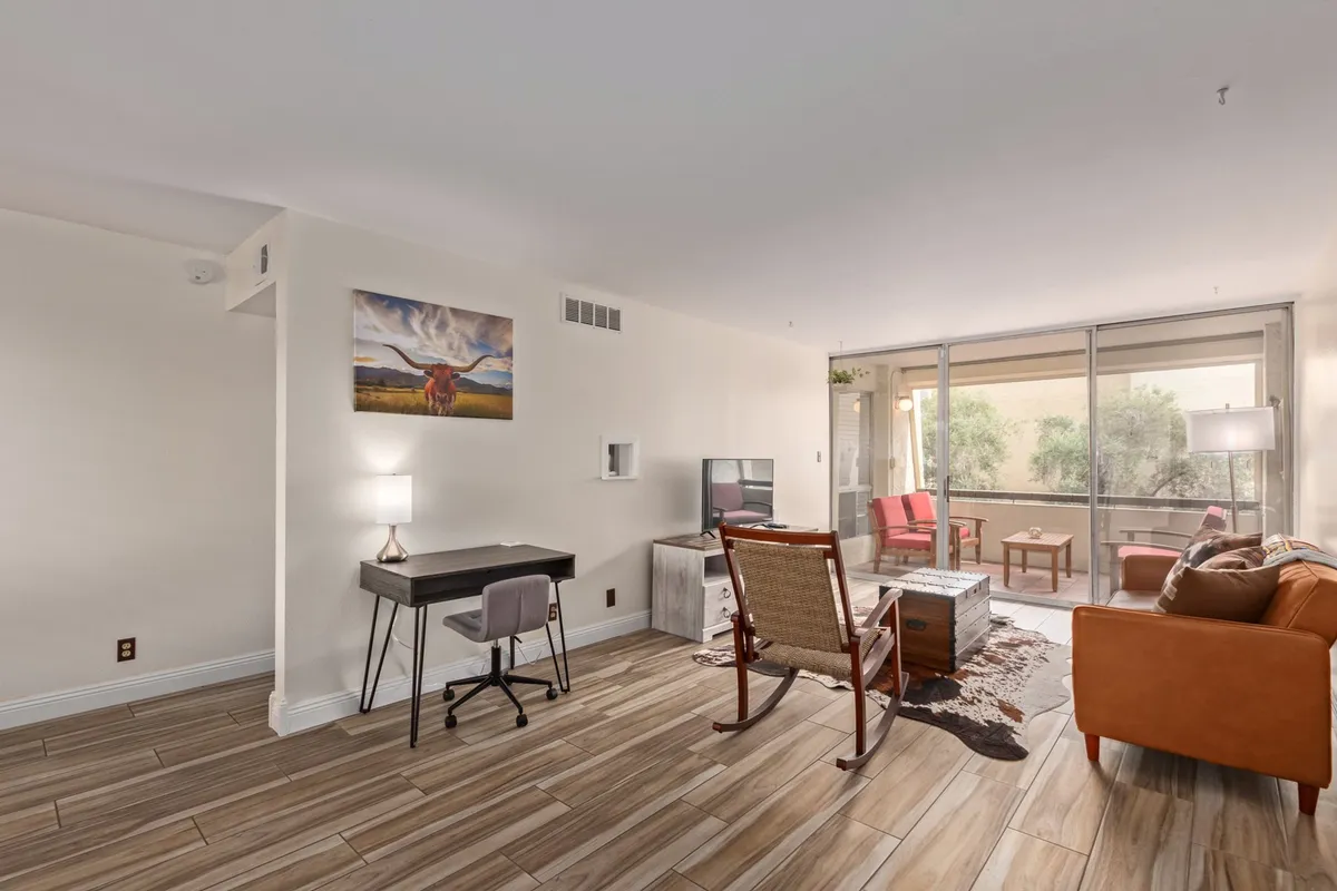 A bright interior room featuring an orange couch, wooden rocking chair, a desk with a lamp, and large windows leading out to the patio or balcony. The windows allow plenty of natural light in and offer a visual connection to the outdoor space.