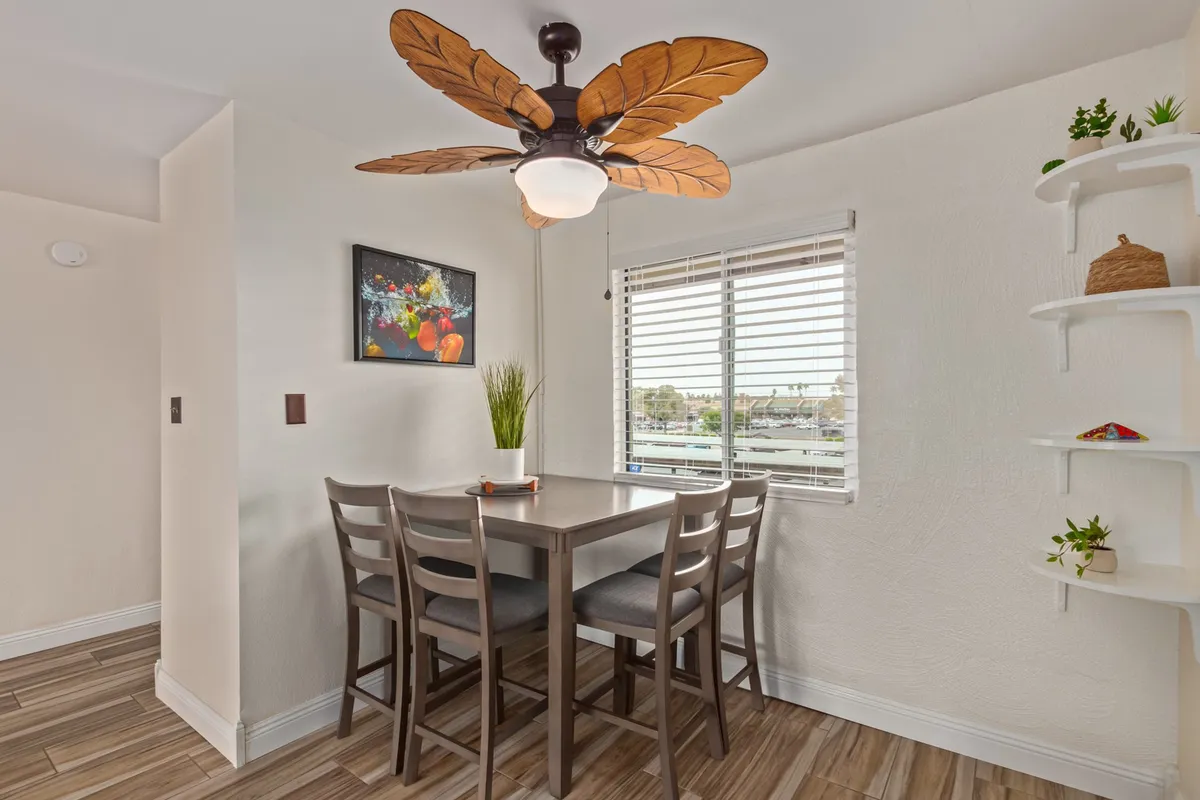 A welcoming dining space featuring a leaf-patterned ceiling fan, a dining table, a large window with blinds, and a stylish decorative shelf. The view through the window hints at the surrounding neighborhood, reinforcing a connection to the outdoors.
