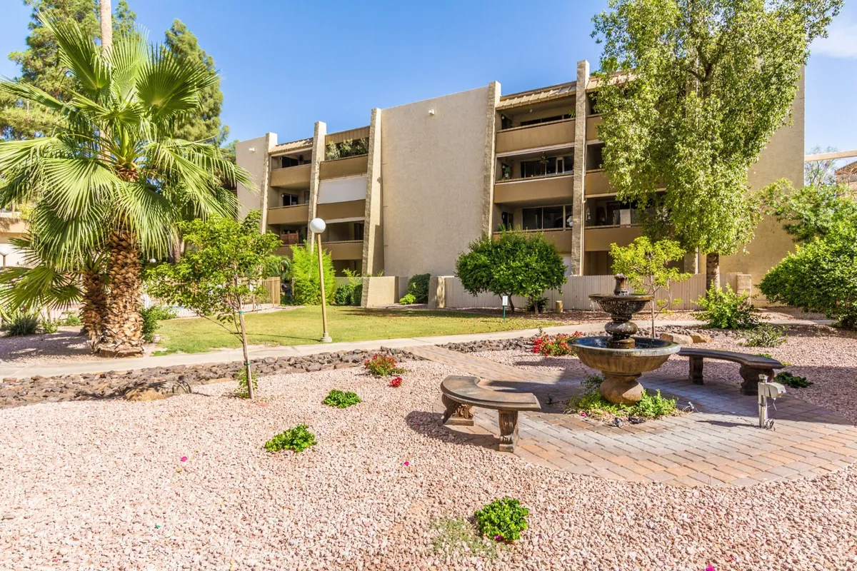 This image features a tranquil outdoor setting with a central fountain surrounded by seating areas, palm trees, and vibrant flower beds. People are seen relaxing by the fountain, socializing, and enjoying the garden atmosphere, with children playing nearby. The scene is lively yet peaceful, reflecting an inviting communal space.