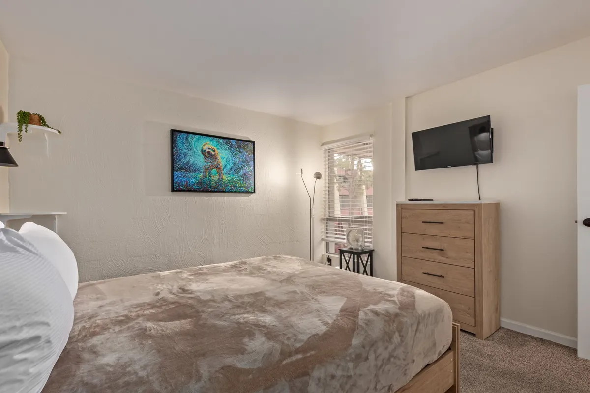 A cozy bedroom featuring a neatly made bed, artwork on the wall, a TV atop a dresser, and a window allowing in natural light. The room exudes comfort and tranquility, suggesting restful mornings and space to unwind.