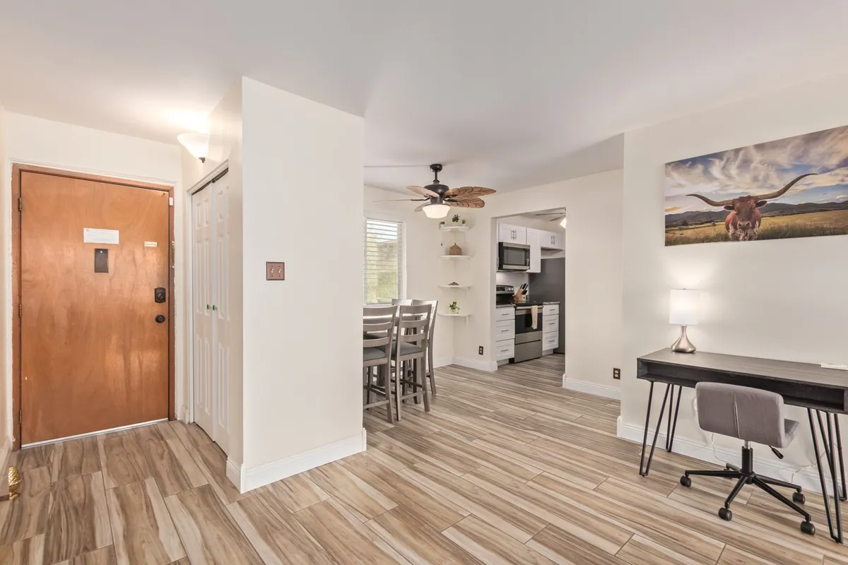 This image shows an inviting open space that includes a front door, dining area, kitchen, wall art, and a convenient desk with chair by the window. The interplay of practical work space with a communal dining area and prominent entryway suggests a dynamic environment ideal for both socializing and everyday routines.