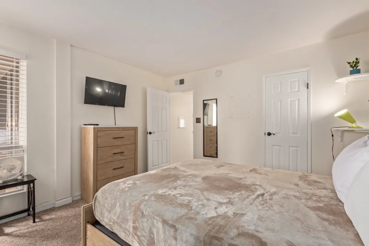 A comfortable bedroom with a large bed, dresser, mirror, and TV, offering plenty of space to rest, get ready, or enjoy a quiet evening. The neutral tones and uncluttered layout enhance the calming atmosphere.