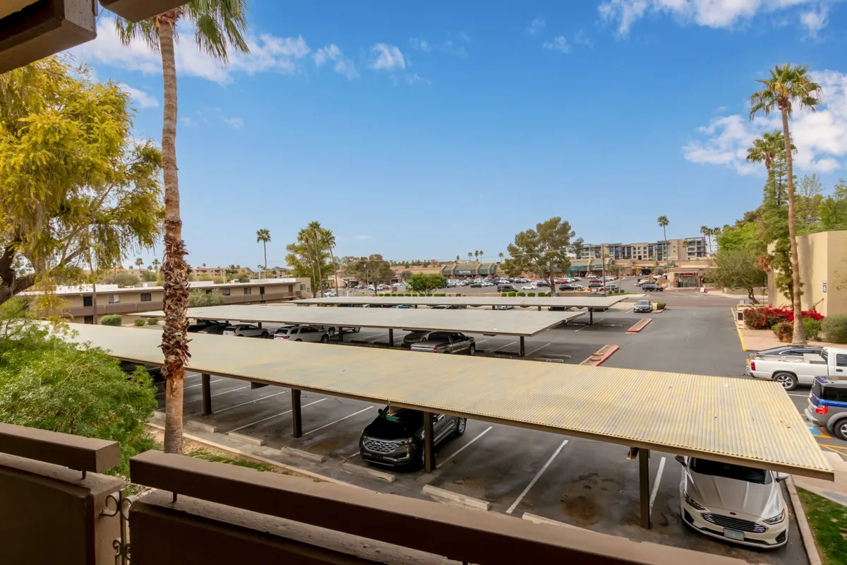 An open-air view of covered parking structures shaded by tall palm trees, with distant buildings suggesting urban proximity. The scene is bathed in sunlight, and the area provides a relaxed transition space from home to the city, inviting residents and visitors to come and go with ease.